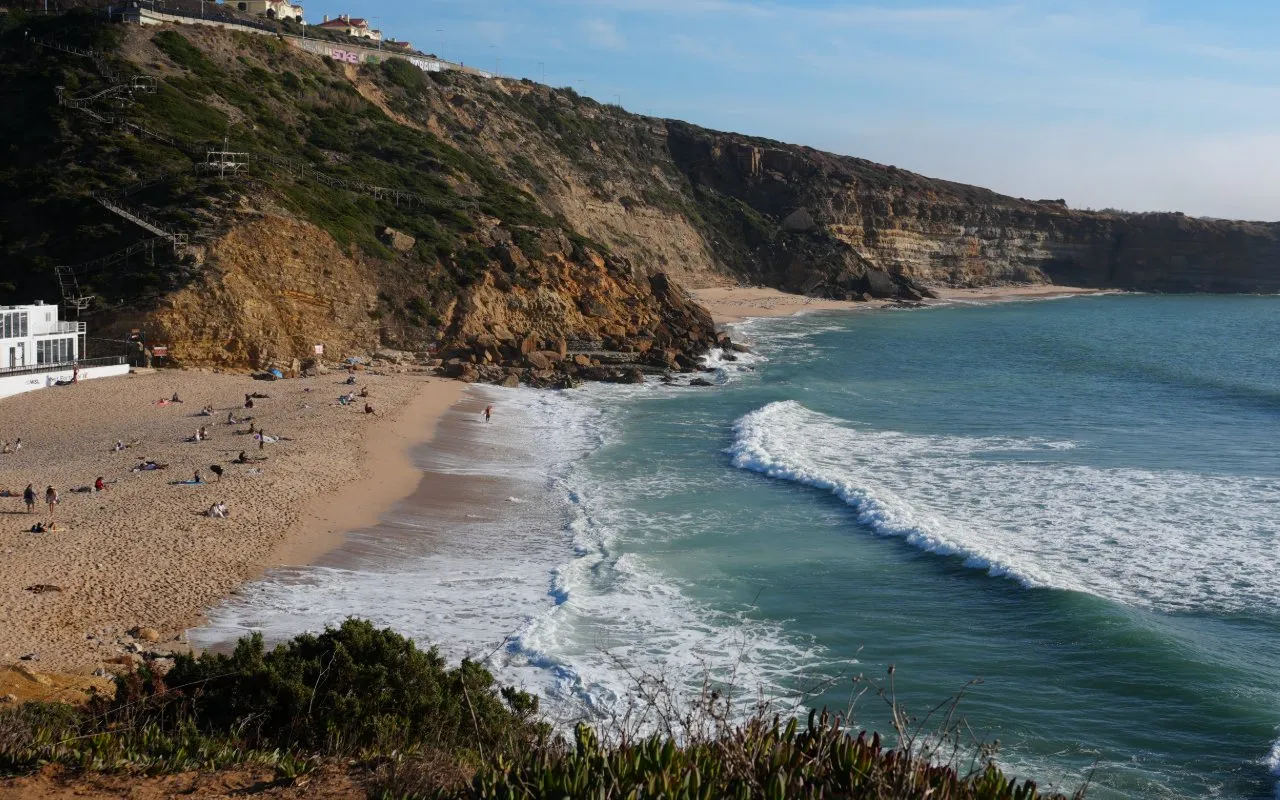 A sandy beach below golden cliffs with waves rolling in on the Ericeira coast