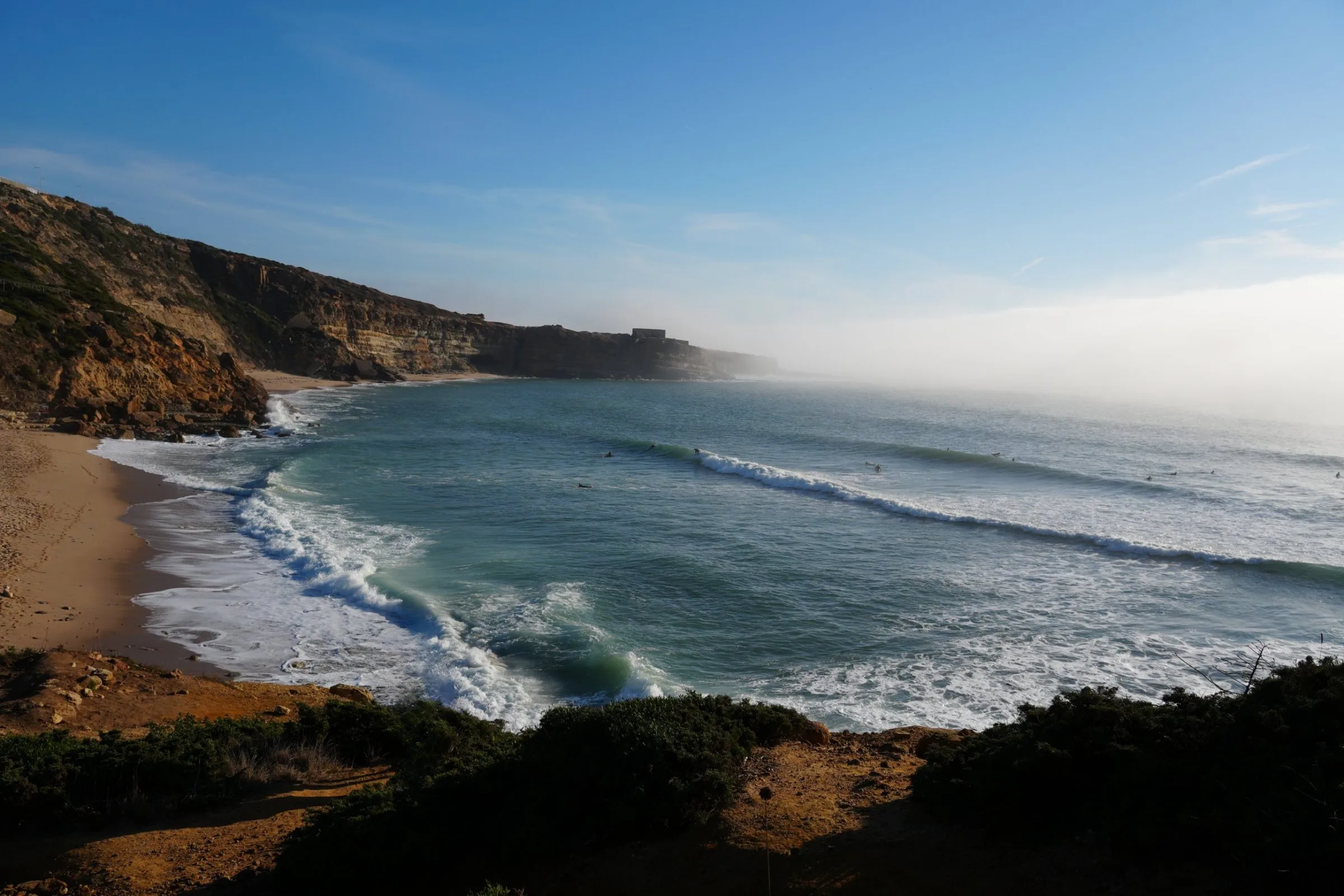 An empty beach curving below the cliffs at Ribeira d'Ilhas, with fog rolling in from the Atlantic